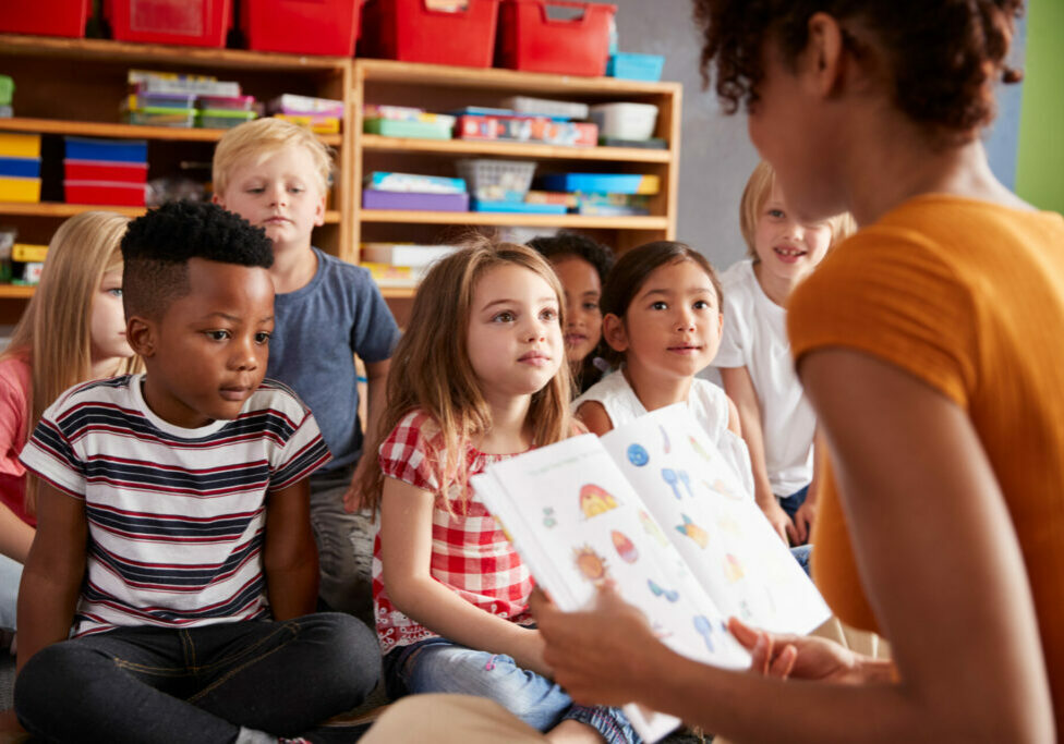 Group Of Elementary School Pupils Sitting On Floor Listening To Female Teacher Read Story